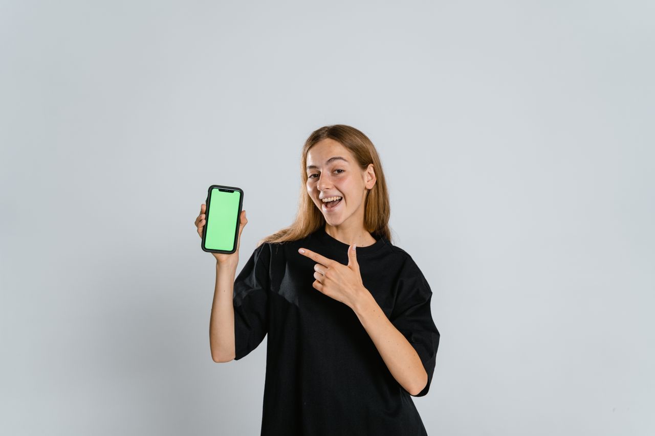 Smiling woman in black t-shirt pointing at a green screen smartphone indoors.